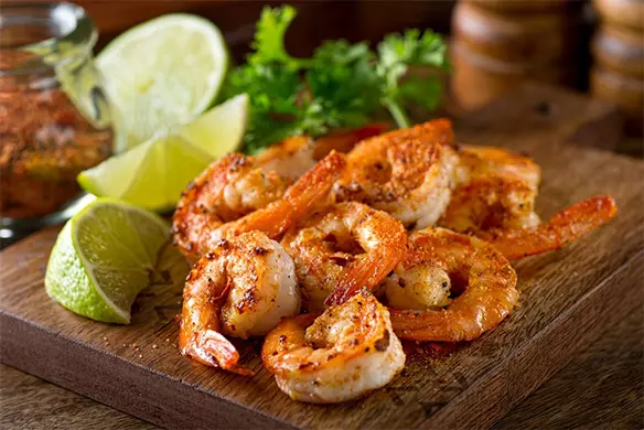 A wooden cutting board topped with shrimp next to lime wedges.